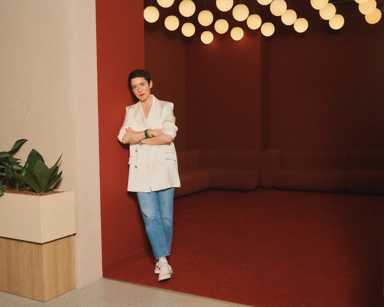 Rachel Delacour, the CEO of Sweep, is leaning against a red wall inside a building. She has short dark brown hair and is wearing a white jacket and jeans.