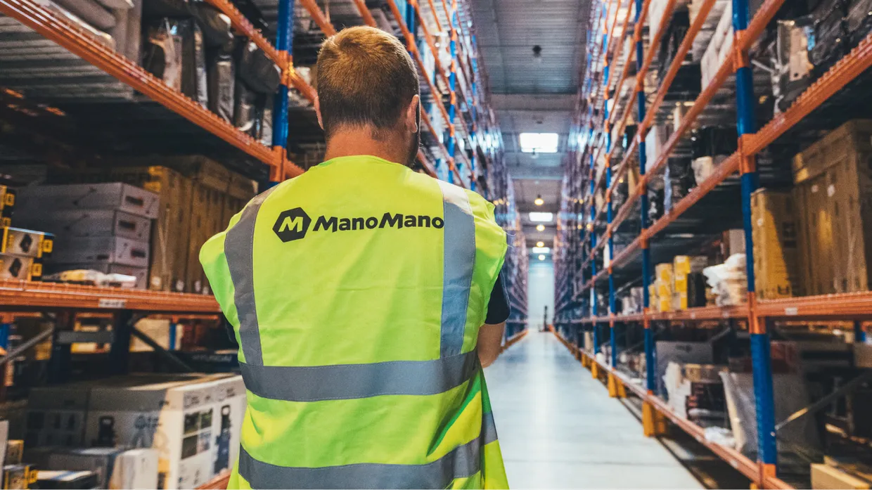 We see the back of a man wearing a hi-vis jacket with the company name "ManoMano". The man is looking into a large warehouse with shelves stacked high with products.